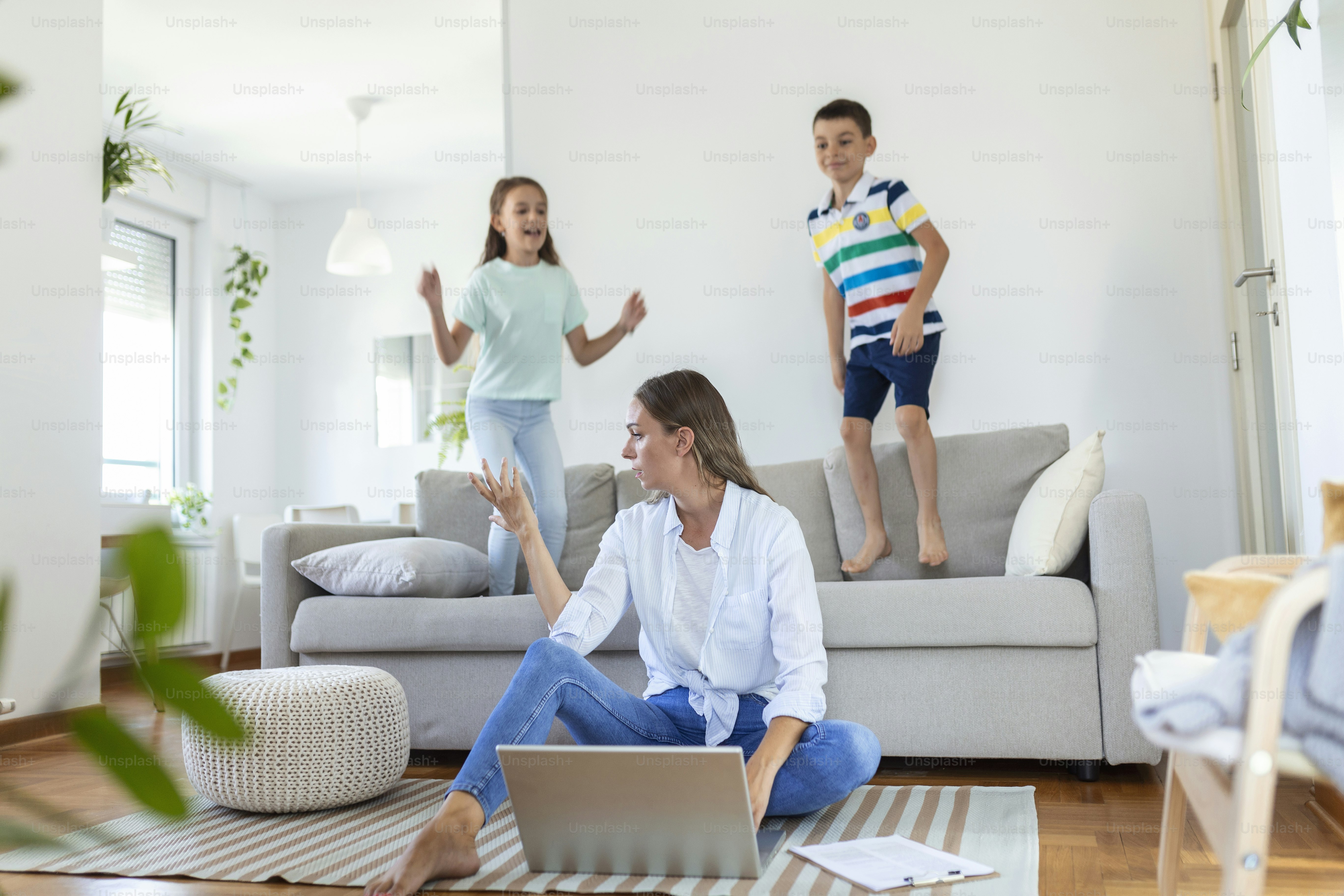 A woman using a laptop on a rug while two children jump on a sofa