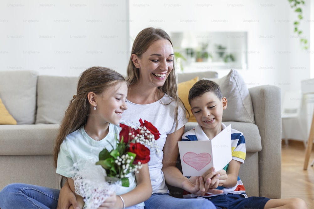 Una madre joven con un ramo de rosas se ríe, abraza a su hijo y una niña con una tarjeta y rosas felicita a su madre durante la celebración de las fiestas en casa. Día de la Madre