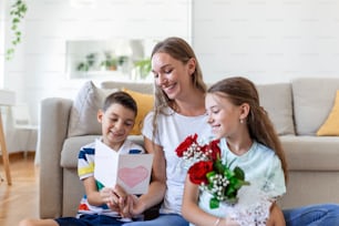 Young mother with a bouquet of roses laughs, hugging her son, and ?heerful girl with a card and roses congratulates mom during holiday celebration at home. mothers day