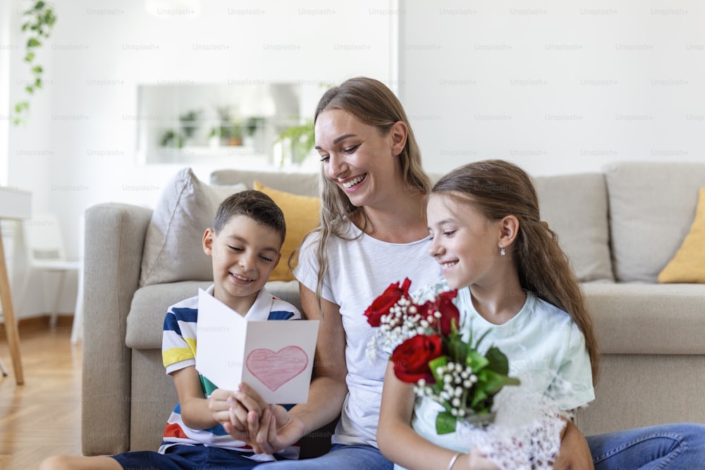 Une jeune mère avec un bouquet de roses rit, étreignant son fils, et une fille avec une carte et des roses félicite maman lors de la fête à la maison. Fête des mères