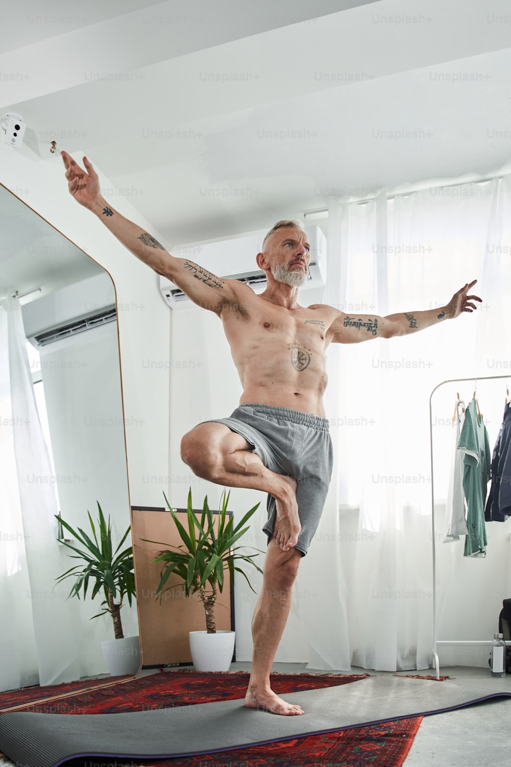 Flachwinkelansicht des ruhigen Mannes mit nacktem Oberkörper, der Yoga in Kriegerpose praktiziert, während er am einen Bein im hellen Wohnzimmer steht. Archivfoto