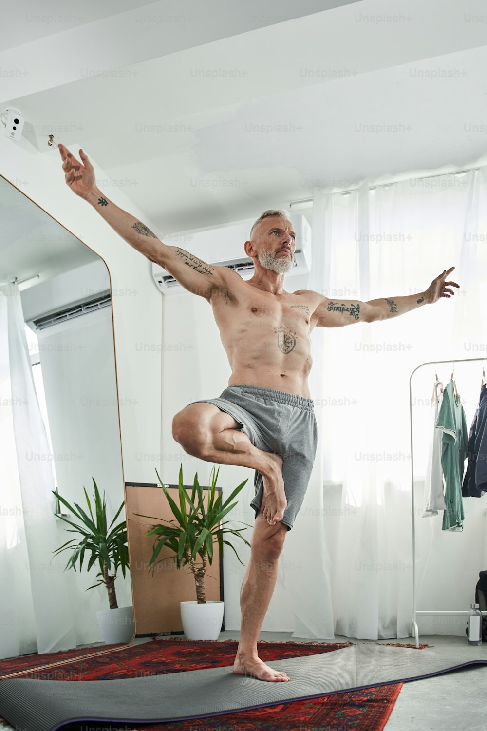 Low angle view of the calm man with naked torso practicing yoga in warrior pose while standing at the one leg at the light living room. Stock photo