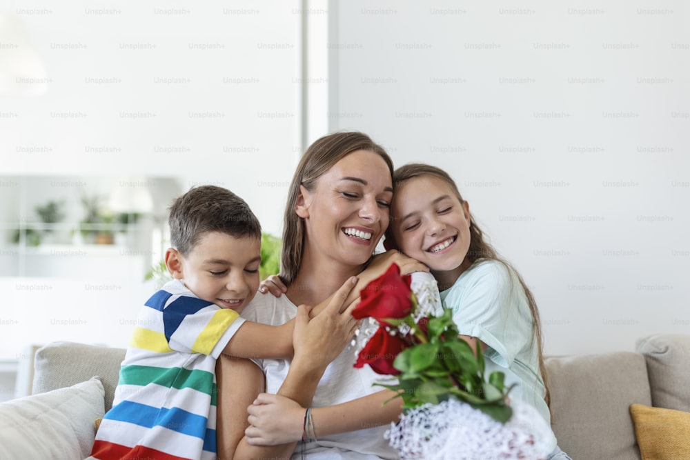 Young mother with a bouquet of roses laughs, hugging her son, and ?heerful girl with a card congratulates mom during holiday celebration in kitchen at home