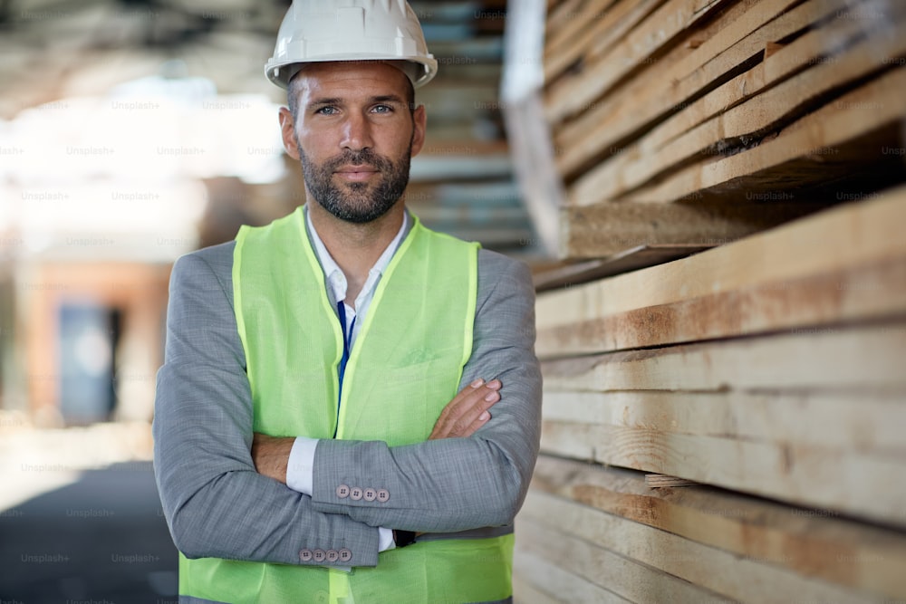 Portrait of businessman standing with crossed arms at lumber distribution warehouse and looking at camera.