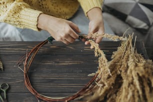 Florist in yellow sweater making rustic autumn wreath on dark wooden table. Hands holding dry grass and making stylish boho wreath with wildflowers and herbs on rustic wooden background.