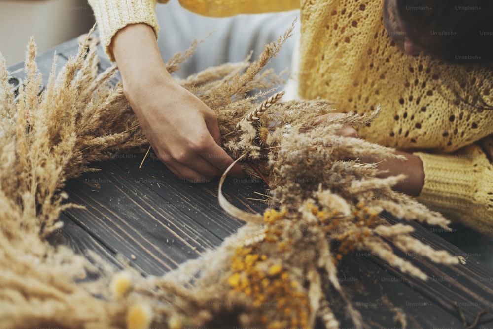 Making stylish autumn wreath. Hands holding dry grass, wildflowers and wheat and making rustic wreath on wooden table. Fall holiday workshop. Florist making boho wreath on dark wood