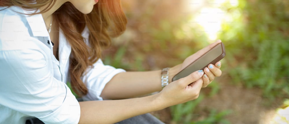 Abgeschnittenes Bild einer Frau, die sich in einem Park entspannt, während sie ihr Smartphone benutzt, um mit ihren Freunden zu kommunizieren. Seitenansicht