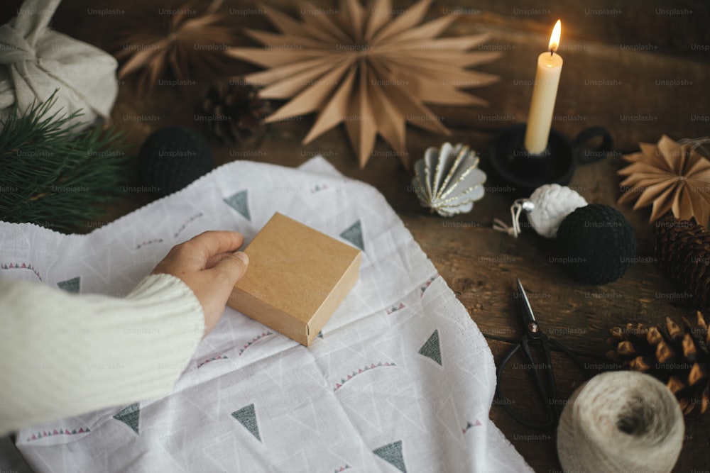 Hands wrapping christmas gift box in modern fabric on rustic wooden table with scissors, craft paper star, candle. Zero waste and eco friendly presents. Atmospheric moody time, nordic style.