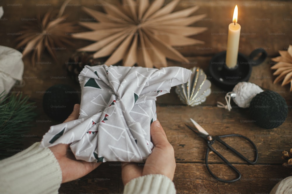Hands holding christmas gift wrapped in modern festive fabric on rustic wooden table with ornaments. Atmospheric moody image, nordic style. Merry Christmas! Furoshiki wrap, zero waste holidays