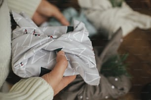 Wrap Furoshiki, vacances de Noël zéro déchet. Mains tenant un cadeau de Noël enveloppé dans un tissu festif moderne sur une table en bois rustique avec des ornements. Image atmosphérique de mauvaise humeur, style nordique