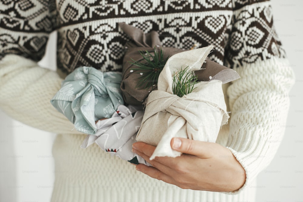 Zero waste and eco friendly presents, Christmas Furoshiki gifts. Woman hands in cozy sweater holding wrapped xmas gift in fabric in scandinavian room. Atmospheric moody image, nordic style.