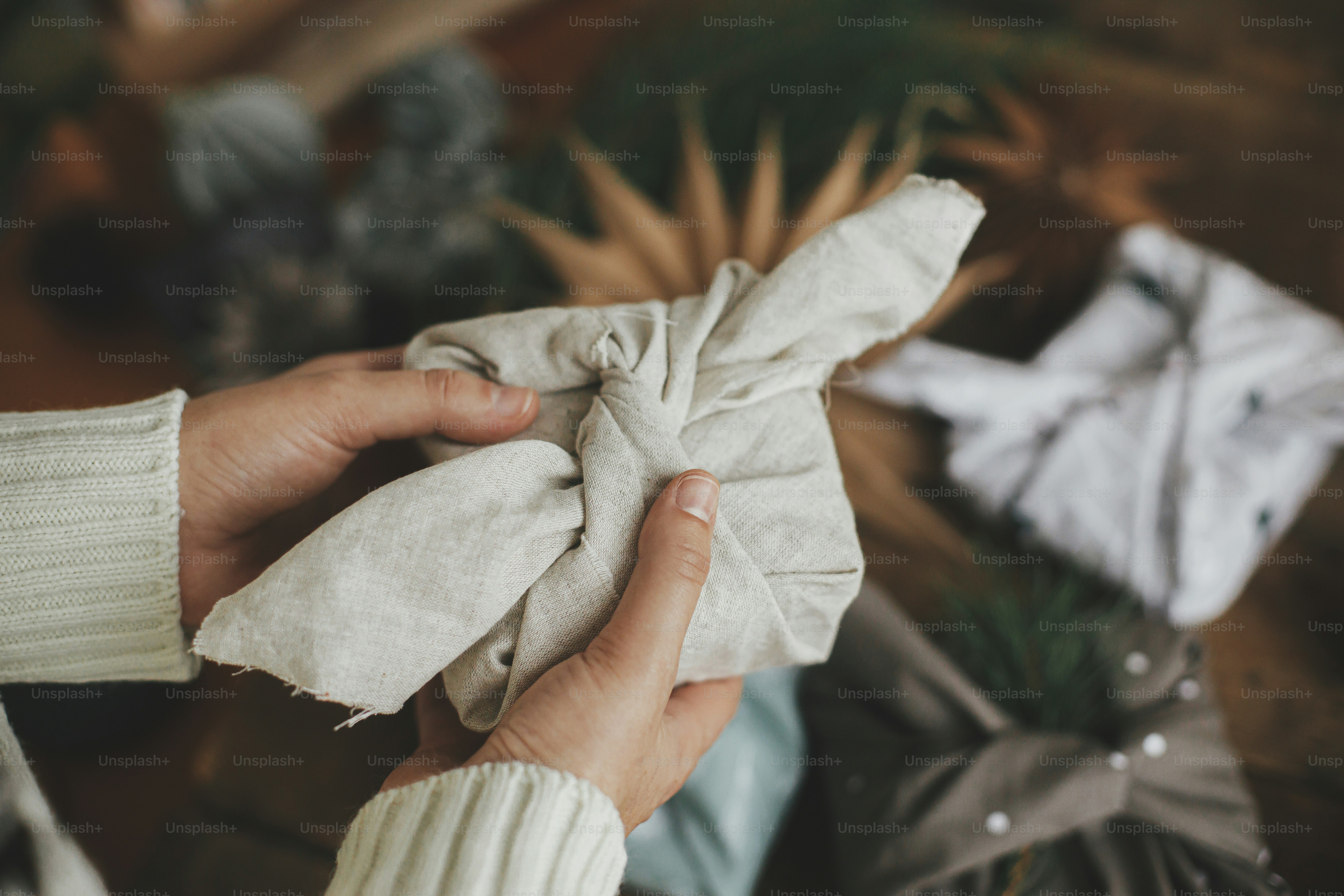 Furoshiki Wrap, Zero Waste Weihnachtsferien. Hände mit Weihnachtsgeschenk verpackt in modernen festlichen Stoff auf rustikalem Holztisch mit Ornamenten. Atmosphärisches stimmungsvolles Bild, nordischer Stil