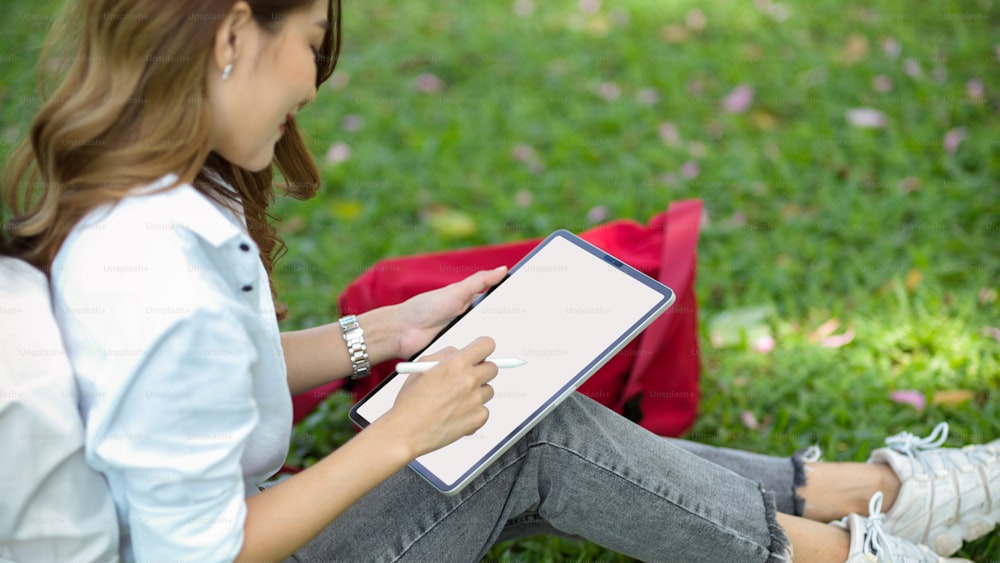 Charming female sits in a park working or sketching picture on digital tablet in a summer day. Tablet blank screen mockup