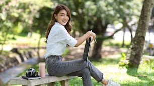 Beautiful female sat on a stone bench with a digital tablet computer, vintage camera, diary, and water bottle, chilling out on an inspiration day in a greenery park.