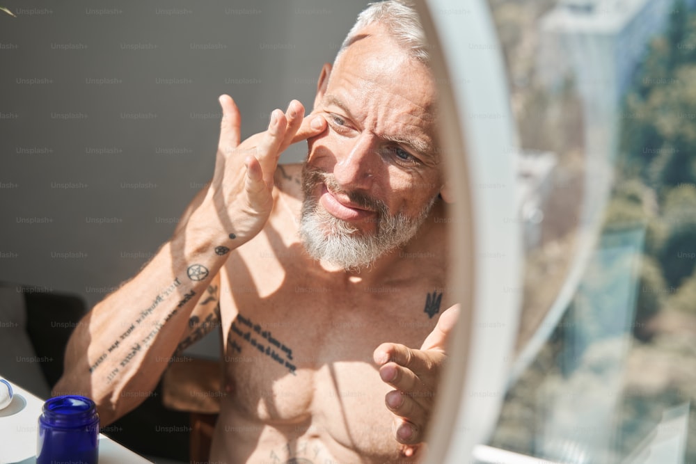 성숙한 남자는 욕실에서 거울을 보면서 얼굴에 노화 방지 크림을 바르고 있다. 얼굴 피부 관리 및 위생의 개념. 국내 생활 방식. 문신을 한 회색 머리 백인 남성 연금 수급자