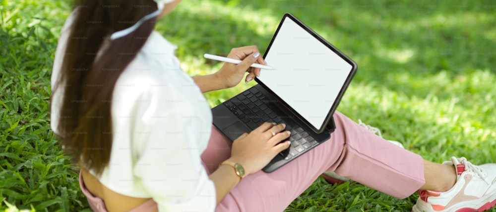 Cropped image of woman sits in a greenery park and works on portable tablet with stylus pen. tablet blank screen mockup