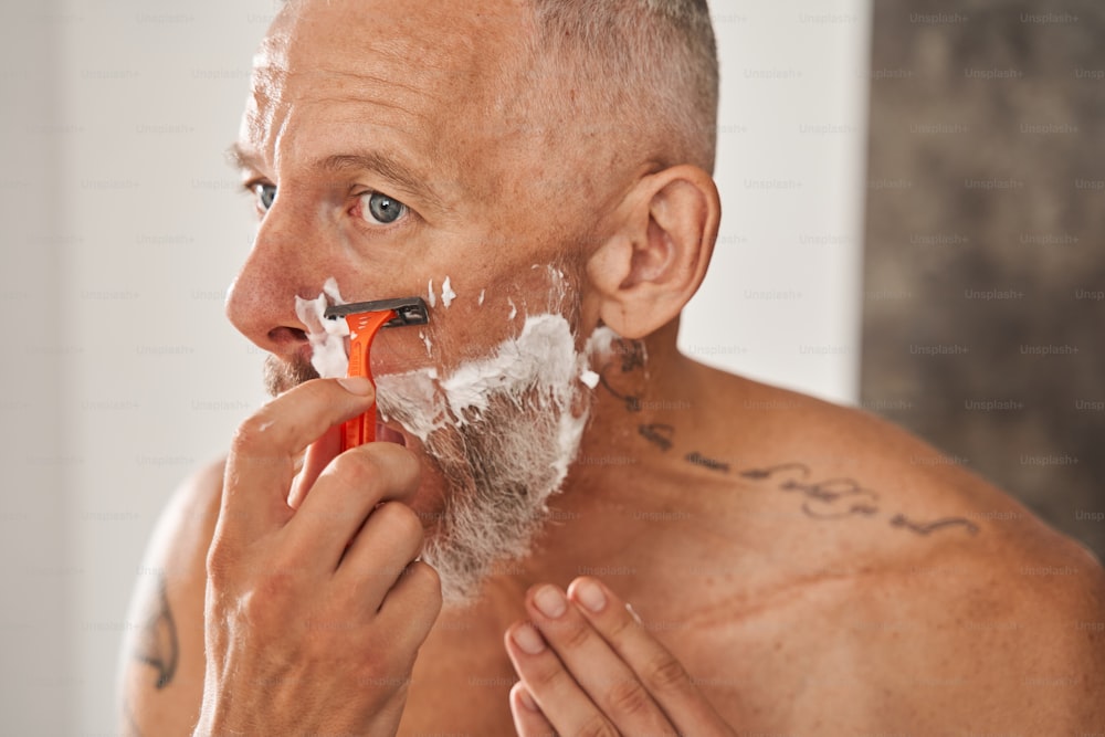 Fast fertig. Porträt eines älteren Mannes mit grauen Haaren, der sich in seinem Schlafzimmer rasiert. Bärtiger Kerl, der Stoppeln loswird. Hautpflegekonzept. Archivfoto
