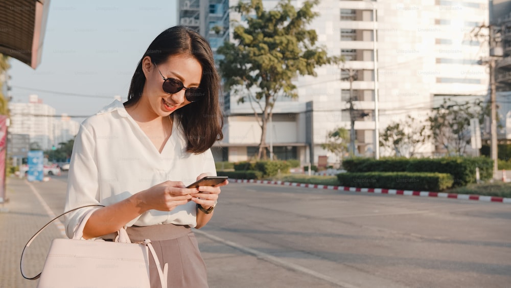 ファッションオフィスの服を着て成功した若いアジアのビジネスウーマンは、都会の近代的な都市の屋外に立って、道路でタクシーを捕まえ、スマートフォンを持っています。外出先でのビジネスのコンセプト。