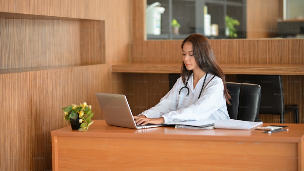 Attraente giovane dottoressa che si concentra sul suo lavoro, scrivendo un documento di ricerca medica sul suo computer portatile.