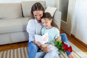 Happy mother's day! Child daughter congratulates mom and gives her postcard and flowers. Mum and girl smiling and hugging. Family holiday and togetherness.