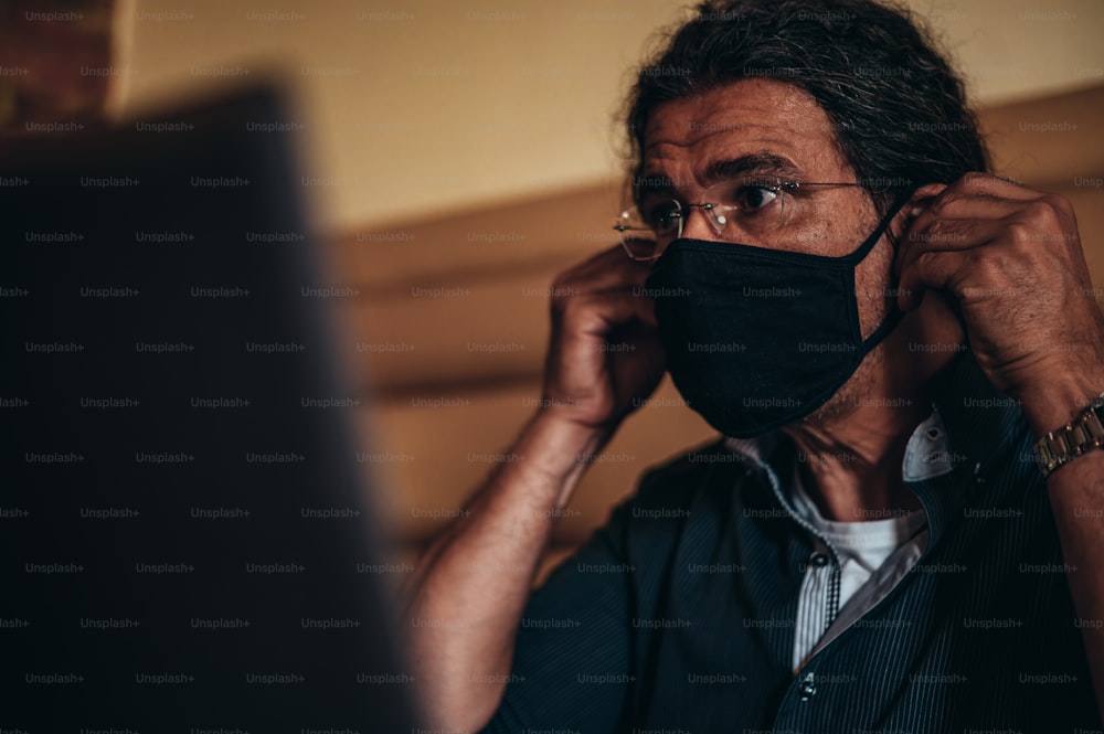カフェに座ってノートパソコンを使いながら保護マスクを着用する年配のヒスパニック系キューバ人男性