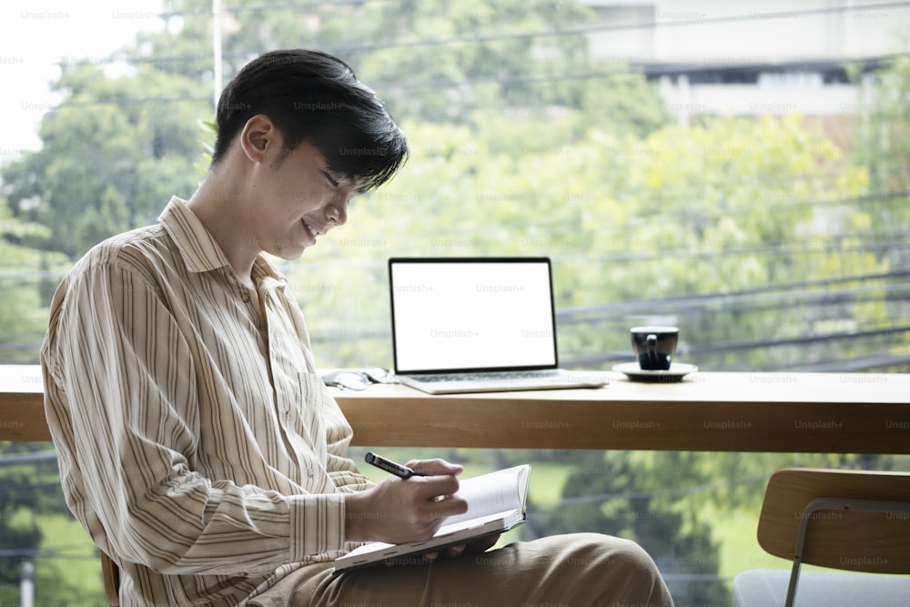 웃고 있는 젊은 아시아 남자가 밝은 사무실에 앉아 노트북에 새로운 아이디어를 쓰고 있다.