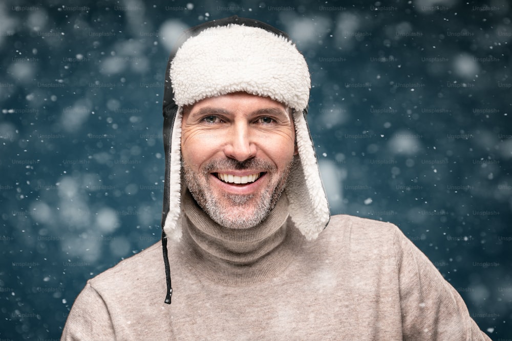 Portrait of handsome man wearing winter cap , posing over snow flakes, looking at camera and smiling. Closeup face of cheerful guy wearing warm clothes.