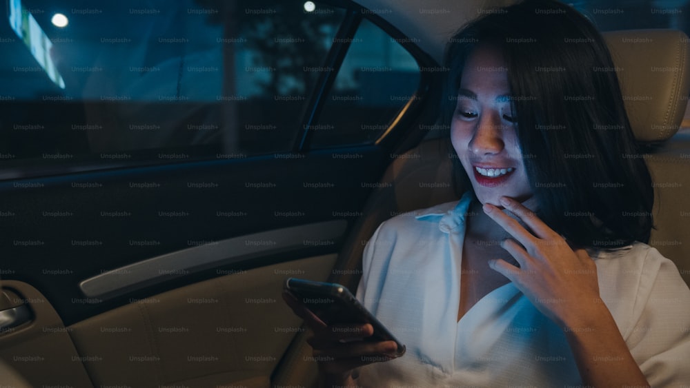 Erfolgreiche junge asiatische Geschäftsfrau in Mode-Bürokleidung arbeitet spät mit Smartphone auf dem Rücksitz des Autos in der urbanen modernen Stadt in der Nacht. Menschen berufliches Burnout-Syndrom Konzept.