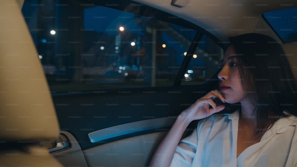 Jeune femme d’affaires asiatique prospère dans les vêtements de bureau de mode travaillant tard à l’aide d’un téléphone intelligent assis sur le siège arrière de la voiture dans une ville urbaine moderne dans la nuit. Concept de syndrome d’épuisement professionnel des personnes.