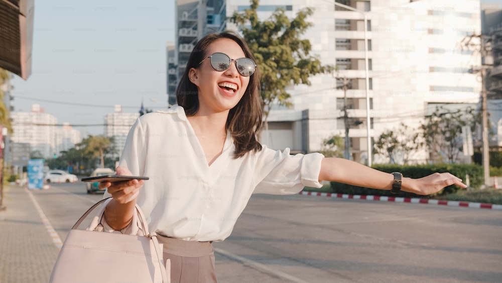 패션 사무복을 입은 성공적인 젊은 아시아 사업가는 택시를 잡고 도시 현대 도시의 야외에 서 있는 동안 스마트폰을 들고 있습니다. 이동 중 비즈니스 개념.