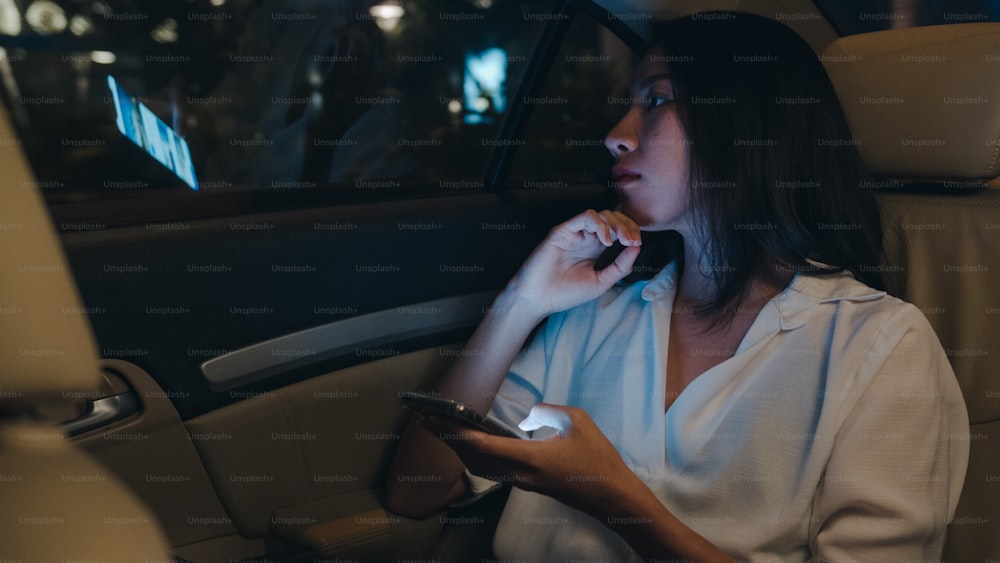 夜の都会の近代的な都市で、車の後部座席に座ってスマートフォンを使って遅くまで働いているファッションオフィス服を着た成功した若いアジアのビジネスウーマン。人々の職業性燃え尽き症候群の概念。