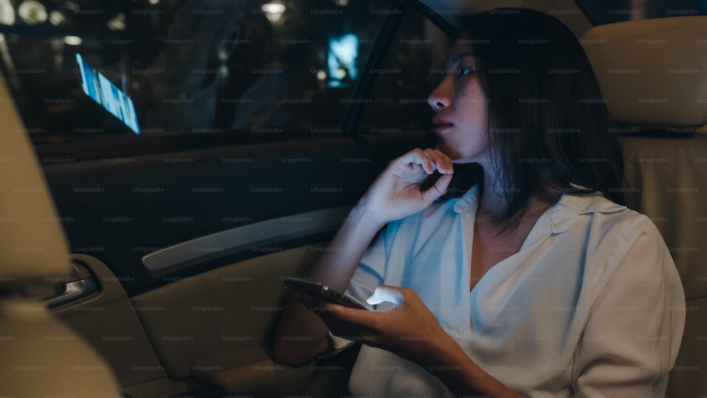 Successful young Asia businesswoman in fashion office clothes working late using smart phone in sitting back seat of car in urban modern city in night. People occupational burnout syndrome concept.