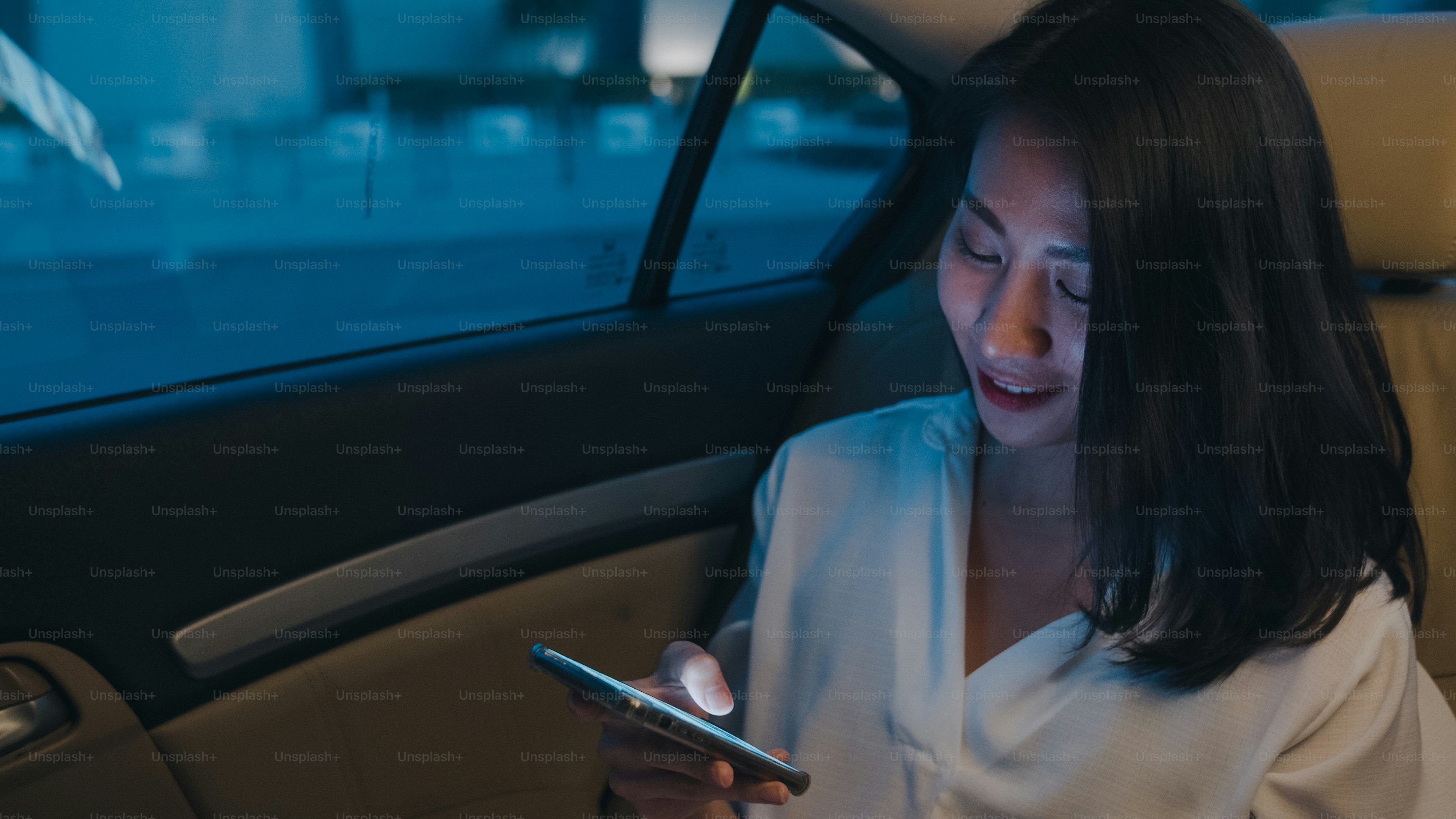 Pengusaha muda Asia yang sukses dalam pakaian kantor fashion bekerja larut malam menggunakan ponsel pintar di kursi belakang mobil di kota modern perkotaan di malam hari. Konsep sindrom kelelahan pekerjaan orang.