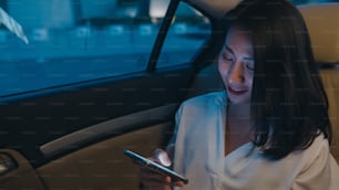 Joven empresaria asiática exitosa en ropa de oficina de moda que trabaja hasta tarde usando un teléfono inteligente en el asiento trasero del automóvil en la ciudad urbana moderna por la noche. Concepto de síndrome de burnout ocupacional de las personas.