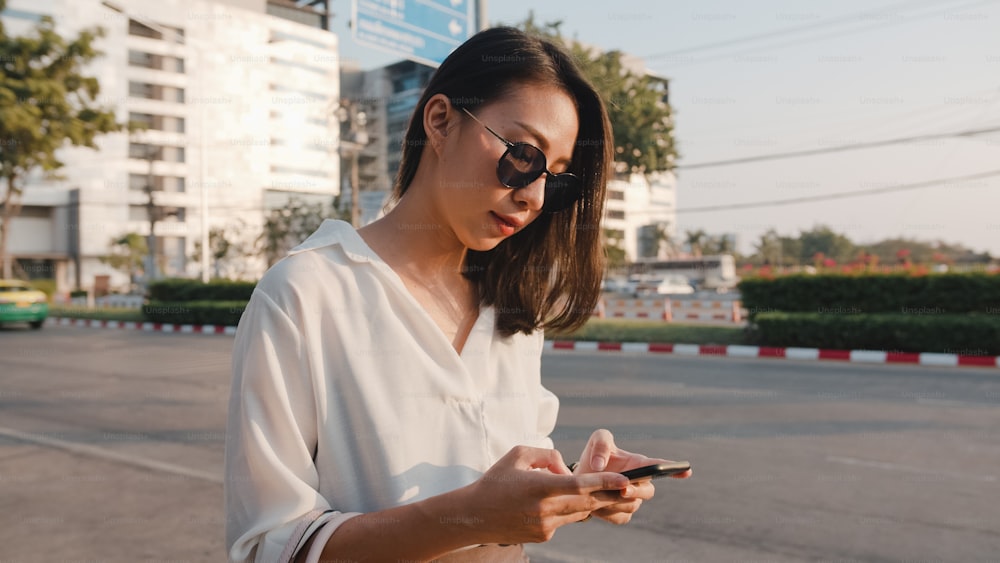 Giovane imprenditrice asiatica di successo in abiti da ufficio di moda che chiama sulla strada che prende il taxi e tiene in mano lo smartphone mentre si trova all'aperto nella città moderna urbana. Concetto di business in movimento.