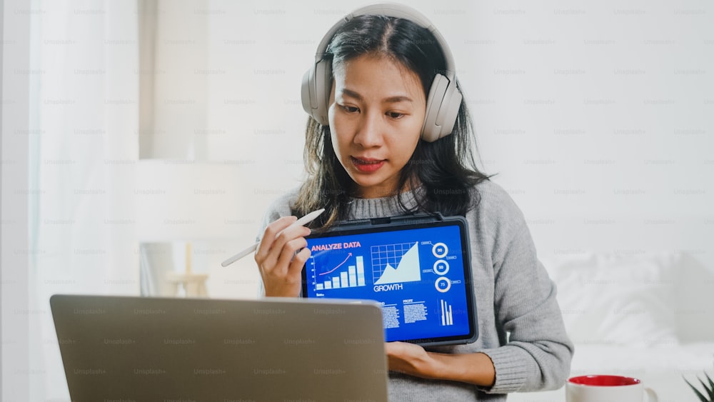 Asia businesswoman wear headphone using laptop talk to colleagues about plan in video call while smart working from home at bedroom. Self-isolation, social distancing, quarantine for corona virus.