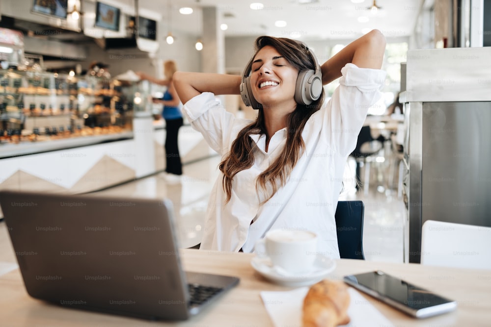 Beautiful and happy young woman sitting and eating delicious rolls in bakery or fast food. She also using her laptop and listening to music with headphones.