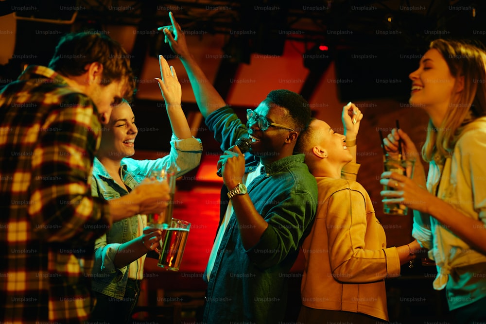 Grupo multiétnico de jóvenes divirtiéndose y cantando karaoke en un bar por la noche. La atención se centra en el hombre negro.