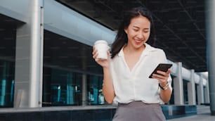 Successful young Asia businesswoman in fashion office clothes holding disposable paper cup of hot drink and using smart phone while standing outdoors in urban modern city. Business on the go concept.