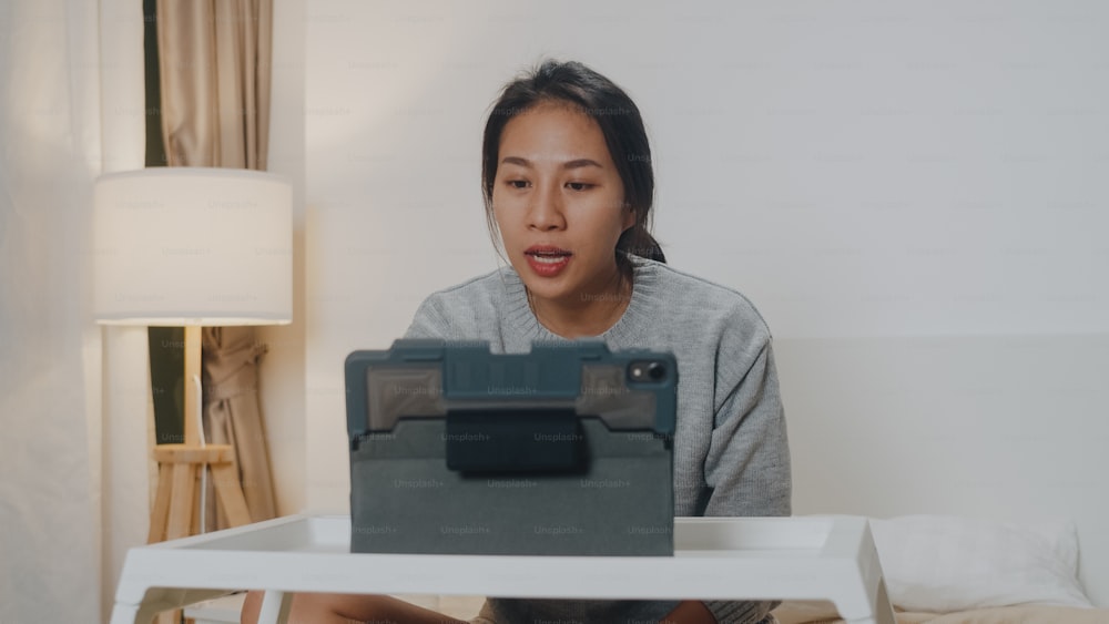 Asia businesswoman using tablet talk to colleagues about plan in video call while smart working from home on bed at bedroom. Self-isolation, social distancing, quarantine for corona virus prevention.