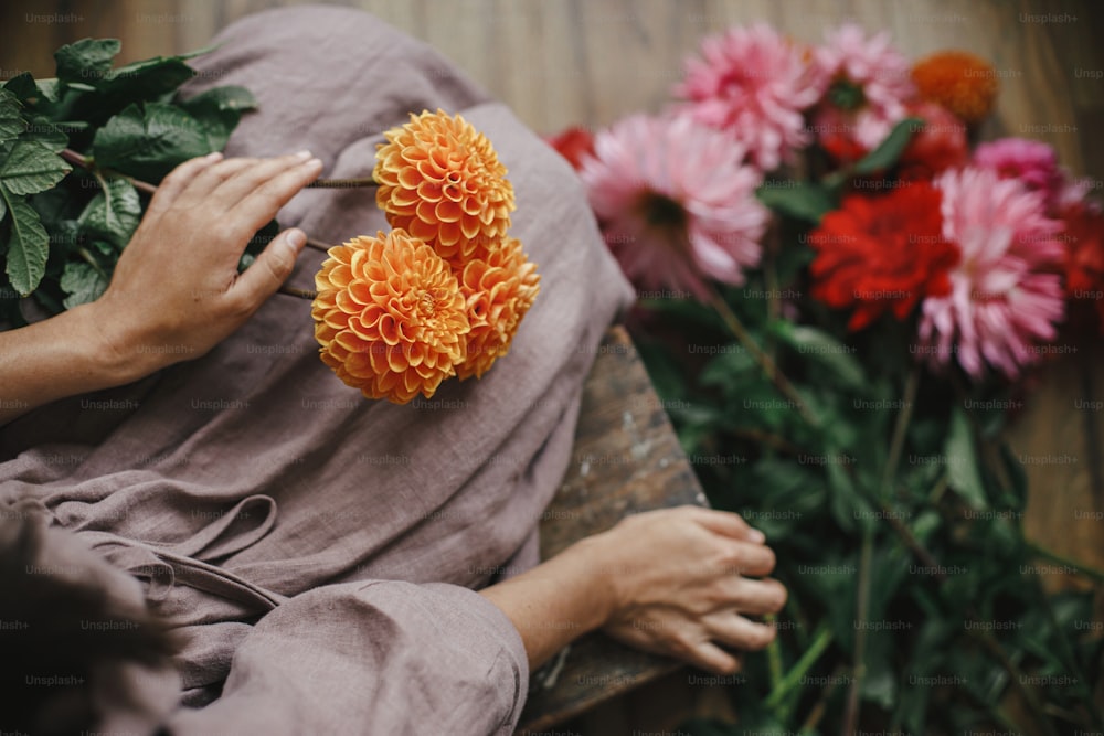 Frau mit orangefarbener Dahlienblüte und auf rustikaler Holzbank sitzend, Blick nach oben. Atmosphärisches stimmungsvolles Bild. Florist im Leinenkleid arrangiert Herbstblumenstrauß auf dem Land. Langsames Leben