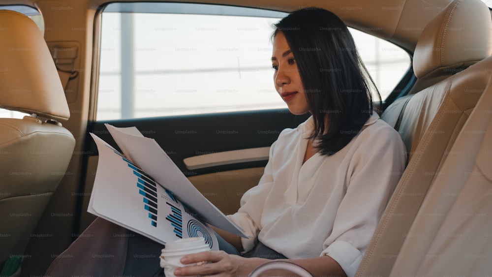 Successful young Asia businesswoman in fashion office clothes working and drinking disposable paper cup of hot drink in back seat of car in urban modern city in morning. Business on the go concept.
