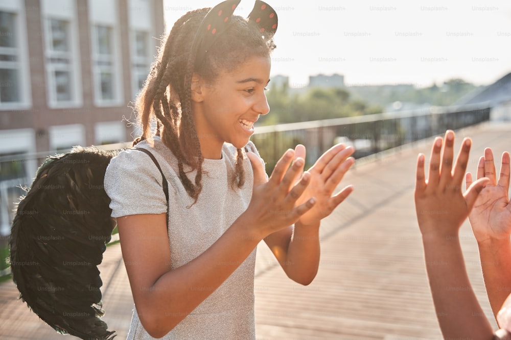 Little black girlfriends playing pat-a-cake on wooden area at halloween. Concept of childhood. Idea of friendship. Modern kid lifestyle. Girl with wings on back and head. Children outdoor at sunny day