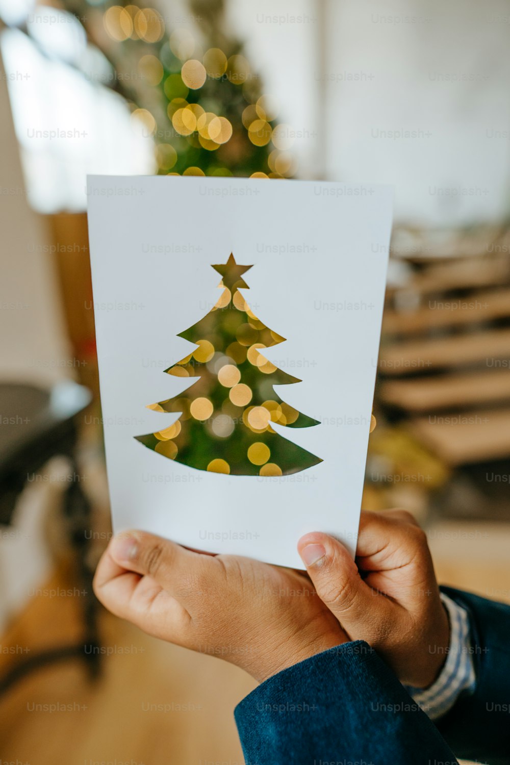Boy holding carved tree on the paper looking throw it at lights on the Christmas tree at home. Idea for DIY handmade festive card.