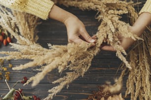 Making rustic autumn boho wreath. Hands holding dry grass and making stylish wreath with wildflowers, herbs and berries on rustic wooden background. Holiday workshop