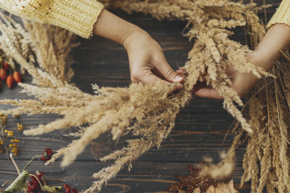 Rustikaler Herbst-Boho-Kranz machen. Hände halten trockenes Gras und machen stilvollen Kranz mit Wildblumen, Kräutern und Beeren auf rustikalem Holzhintergrund. Ferienwerkstatt