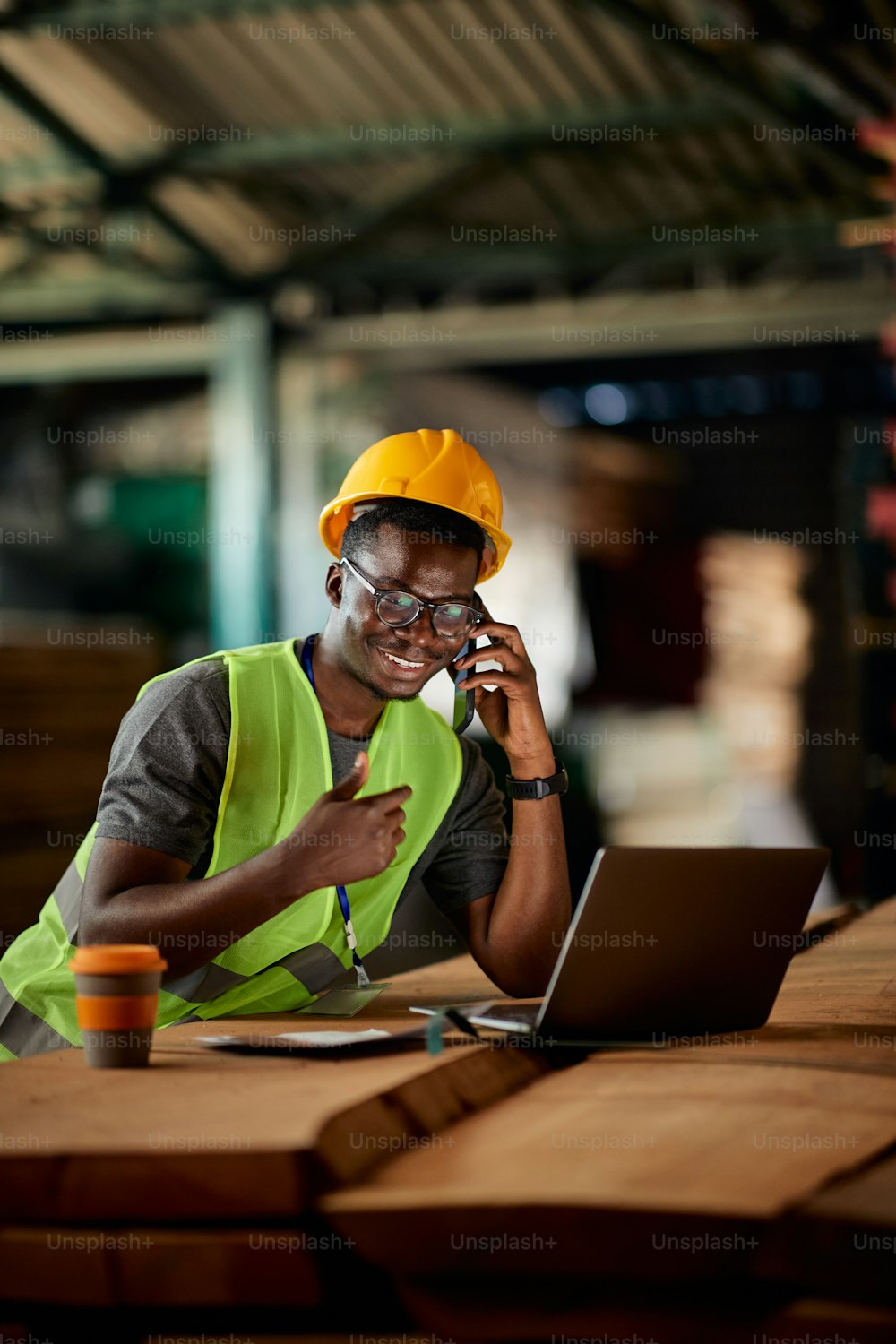 Happy African American warehouse worker communicating over mobile phone while working on laptop.