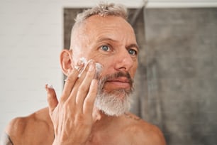 Preparing for new coming day. Waist up portrait of happy mature man applying shaving foam on his face while standing in bathroom before mirror