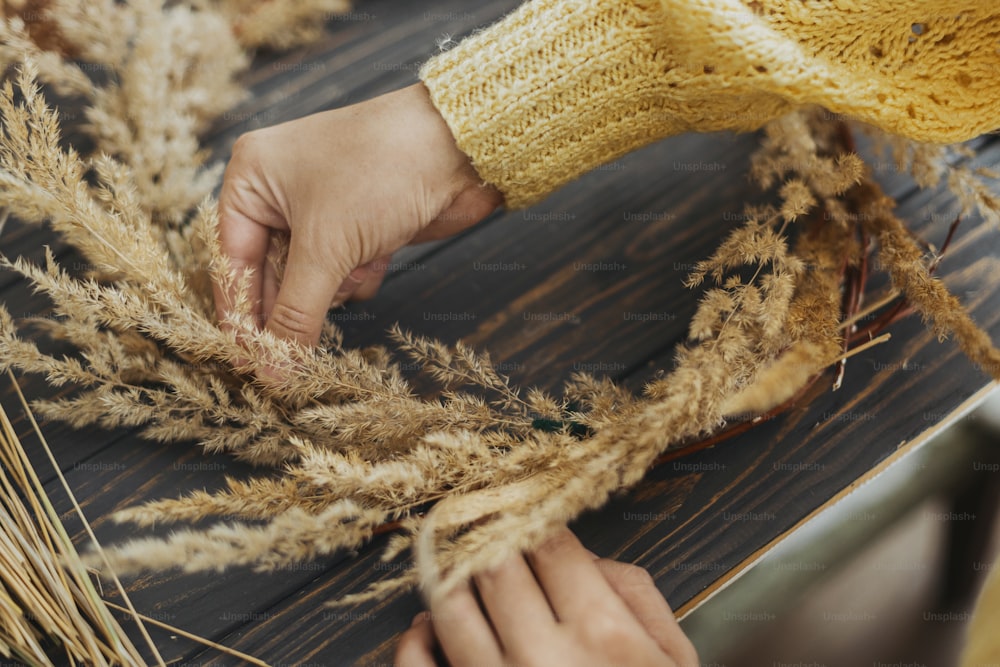 Florist in yellow sweater making rustic autumn wreath on dark wooden table. Hands holding dry grass and making stylish boho wreath with wildflowers and herbs on rustic wooden background.