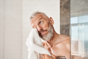 Vista de retrato do homem barbudo maduro limpando o rosto com toalha branca pela manhã. Masculino com conceito de rotina de beleza diária